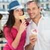 Portrait of a smiling couple eating ice cream in the city. The grey hair man with a beard is in a white shirt. The woman is wearing a blue hat and a pink top.